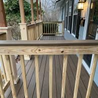 A wooden porch railing with natural-toned balusters and a dark brown top rail, overlooking a deck and a house exterior.