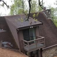 An elevated view of a brown shingled roof featuring a small balcony and a stone chimney under a tree branch.