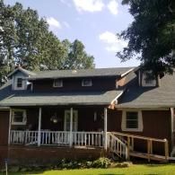 A two-story brown house with a green roof, a front porch with white railings, and a wooden wheelchair ramp.