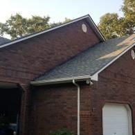 A view of a brown brick house exterior with dark shingled roofs, white gutters, and a garage door under a clear sky.