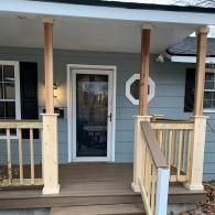 A front porch with a gray-sided house, light-colored wooden railings, and pillars leading to a glass-paneled front door.