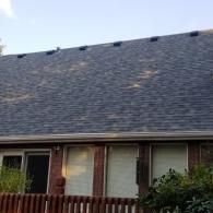 Exterior view of a brick house with a dark shingled roof, a row of roof vents, and a wooden fence in the foreground.