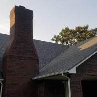 A brown brick chimney stands beside a house roof with dark shingles, white gutters, and a tree against a clear sky.