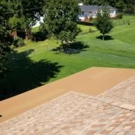 A high-angle view of a beige rooftop and flat deck overlooking a green, grassy backyard with trees and a distant house.