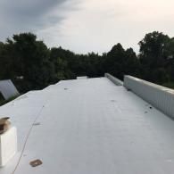A wide shot of a flat, light-gray commercial roof under a cloudy sky, bordered by a metal parapet wall and distant trees.