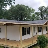 A white house with a newly constructed wooden wrap-around porch and a fresh gray shingled roof under a cloudy sky.
