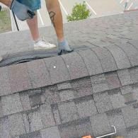 A person wearing gloves and a shoe stands on a shingled roof, holding a tool while working on the ridge cap shingles.