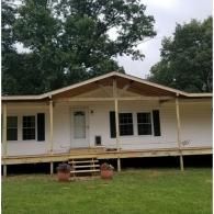 A light-colored single-story home with a new wooden porch and steps in front of a tree-lined yard under a cloudy sky.