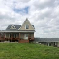 A tan, two-story house with a prominent A-frame gable, wrap-around porch, and an attached side garage under a cloudy sky.