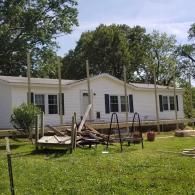 A white, single-story mobile home under renovation with vertical support beams and construction debris in the front yard.