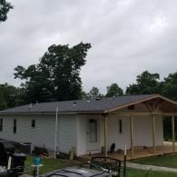 A single-story white house with light gray vinyl siding and a newly constructed, unfinished wooden porch under a gray sky.