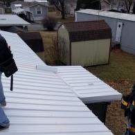 A white metal roof with a partially installed ridge cap, viewed from above, with a small shed in the background.