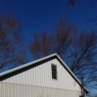 Gable end of a white metal-sided building with a single window under a clear, bright blue sky and bare tree branches.