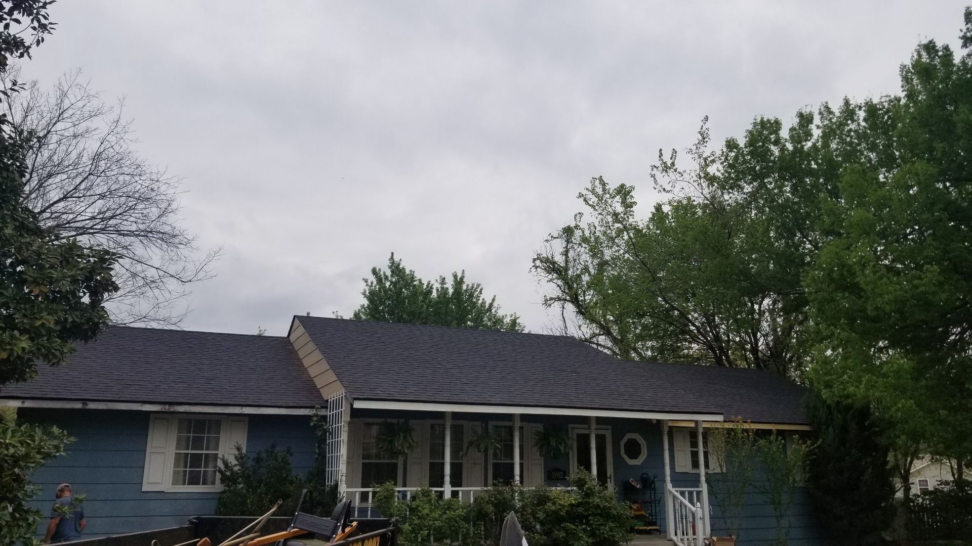 A blue house with a dark gray shingled roof, a covered front porch, and surrounding green trees under a cloudy sky.