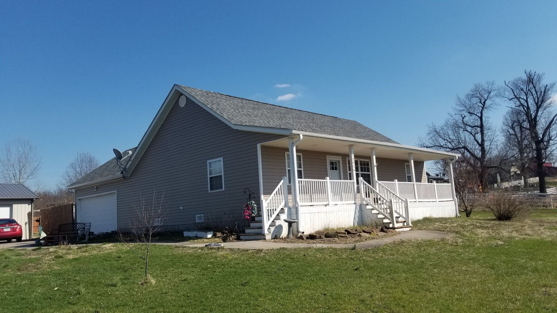 A gray, single-story house with a white front porch and attached garage on a grassy lot under a clear blue sky.