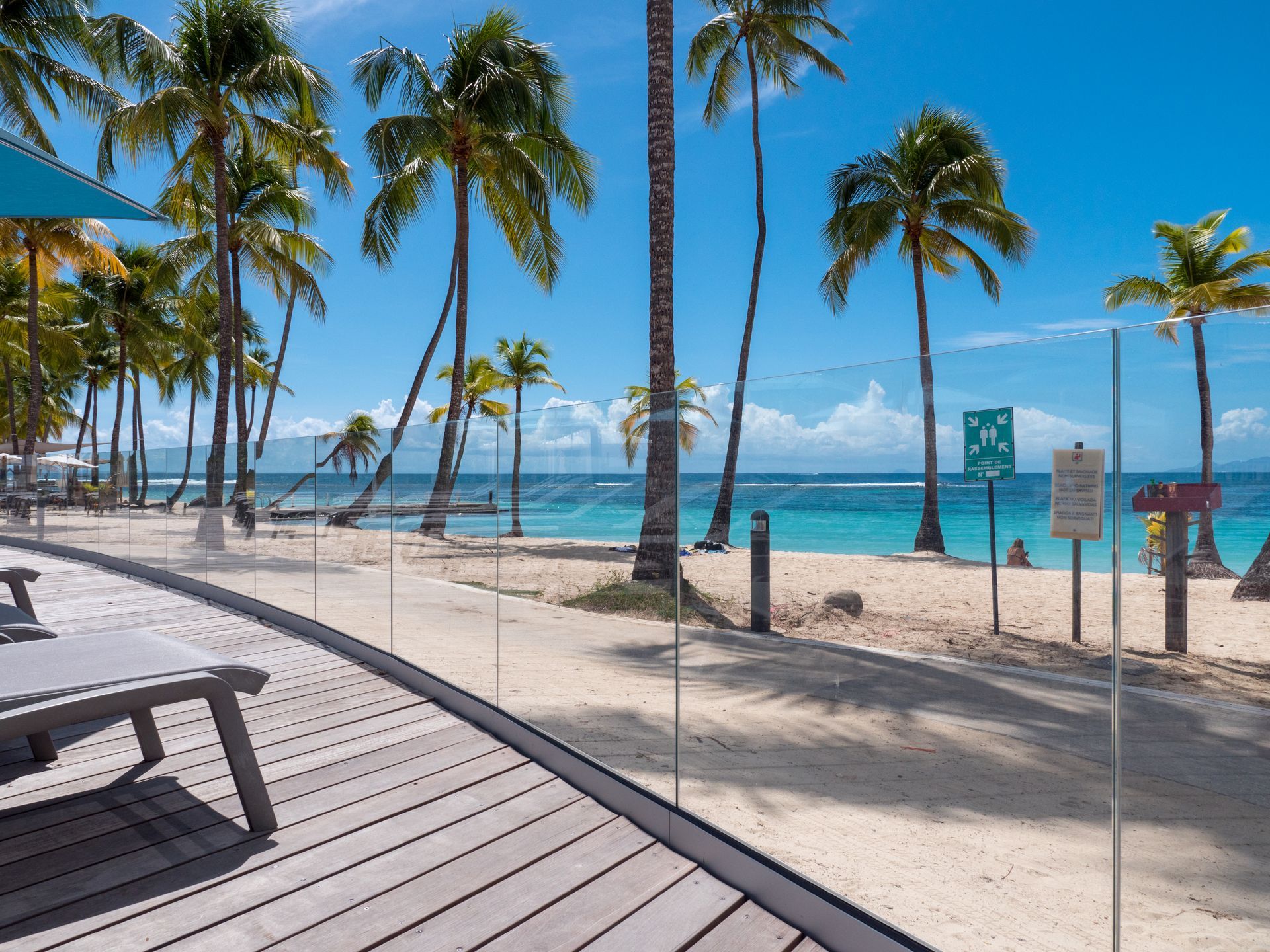 Scène de plage avec palmiers, eau turquoise et terrasse en bois.