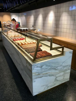 Vitrine remplie de pâtisseries dans une boulangerie. Comptoir en marbre blanc, garnitures dorées et personne en arrière-plan.