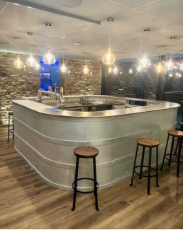 Bar avec comptoir argenté, tabourets, mur en briques apparentes, luminaires suspendus.