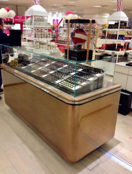 Présentoir de chocolats dans un magasin, présentant diverses gourmandises sous verre. Bois brun clair et design moderne.