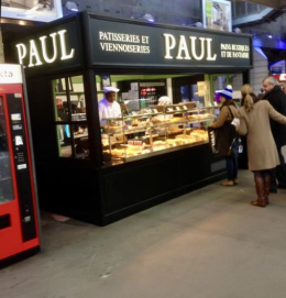 Un stand de boulangerie Paul avec des clients. Façade noire et blanche, pâtisseries exposées. Des clients achètent à manger.