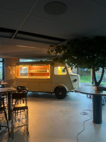 Un food truck, beige avec des lumières chaudes, est installé à l'intérieur d'un bâtiment près d'un arbre et de sièges.