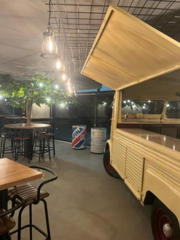 Un bar intérieur/extérieur avec un food truck vintage, des tables, des tabourets, des guirlandes lumineuses et un arbre.