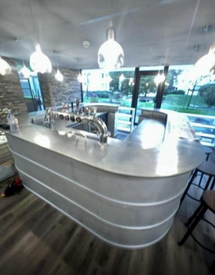 Bar avec robinets argentés, béton gris courbé, luminaires suspendus et vue sur la verdure.