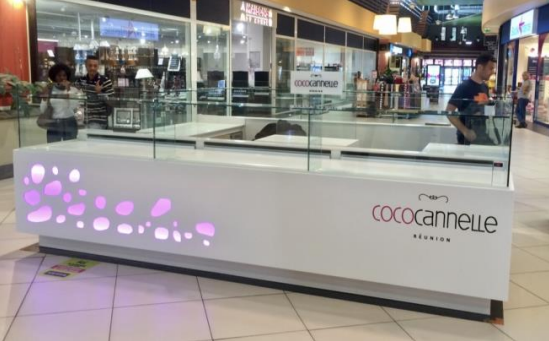 Un kiosque blanc Coco Cannelle dans un centre commercial avec du personnel et un présentoir, éclairé en violet.