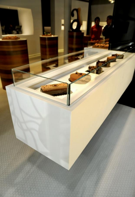 Vitrine blanche avec des pâtisseries, dans un décor de boulangerie ; deux personnes visibles en arrière-plan.