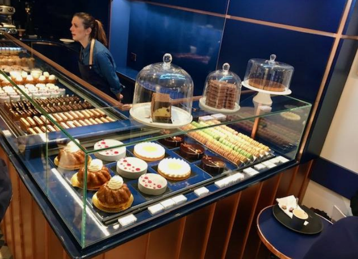 Une vitrine de pâtisserie remplie de gâteaux, de tartes et de macarons. Une femme se tient derrière le comptoir.