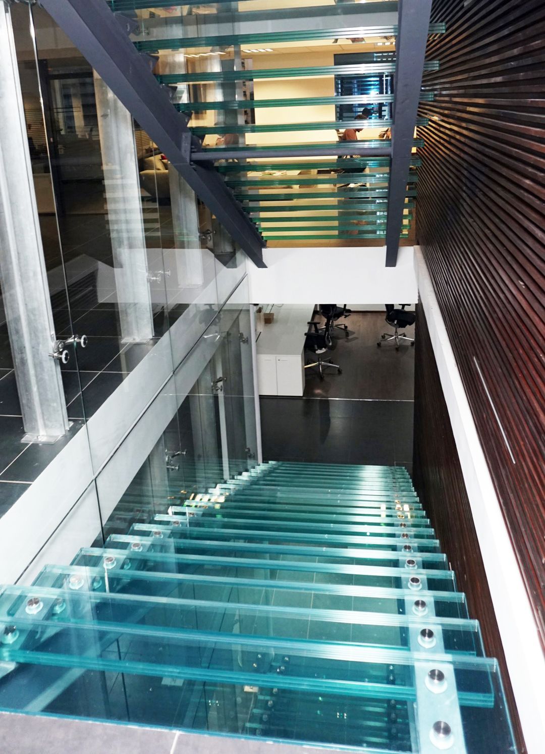 Escalier en verre vu d'en haut, descendant vers un sol sombre. Mur en bois à droite.