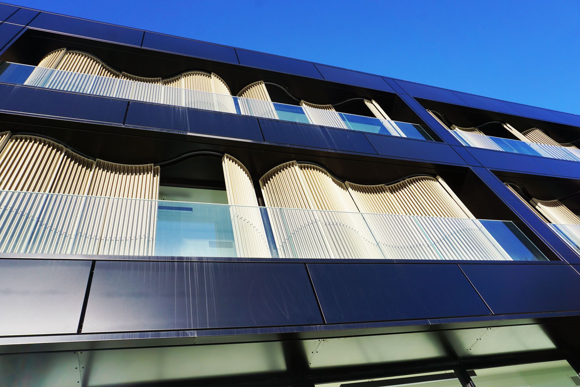 Façade d'un bâtiment moderne avec panneaux sombres et parasols ondulés couleur crème. Ciel bleu en arrière-plan.