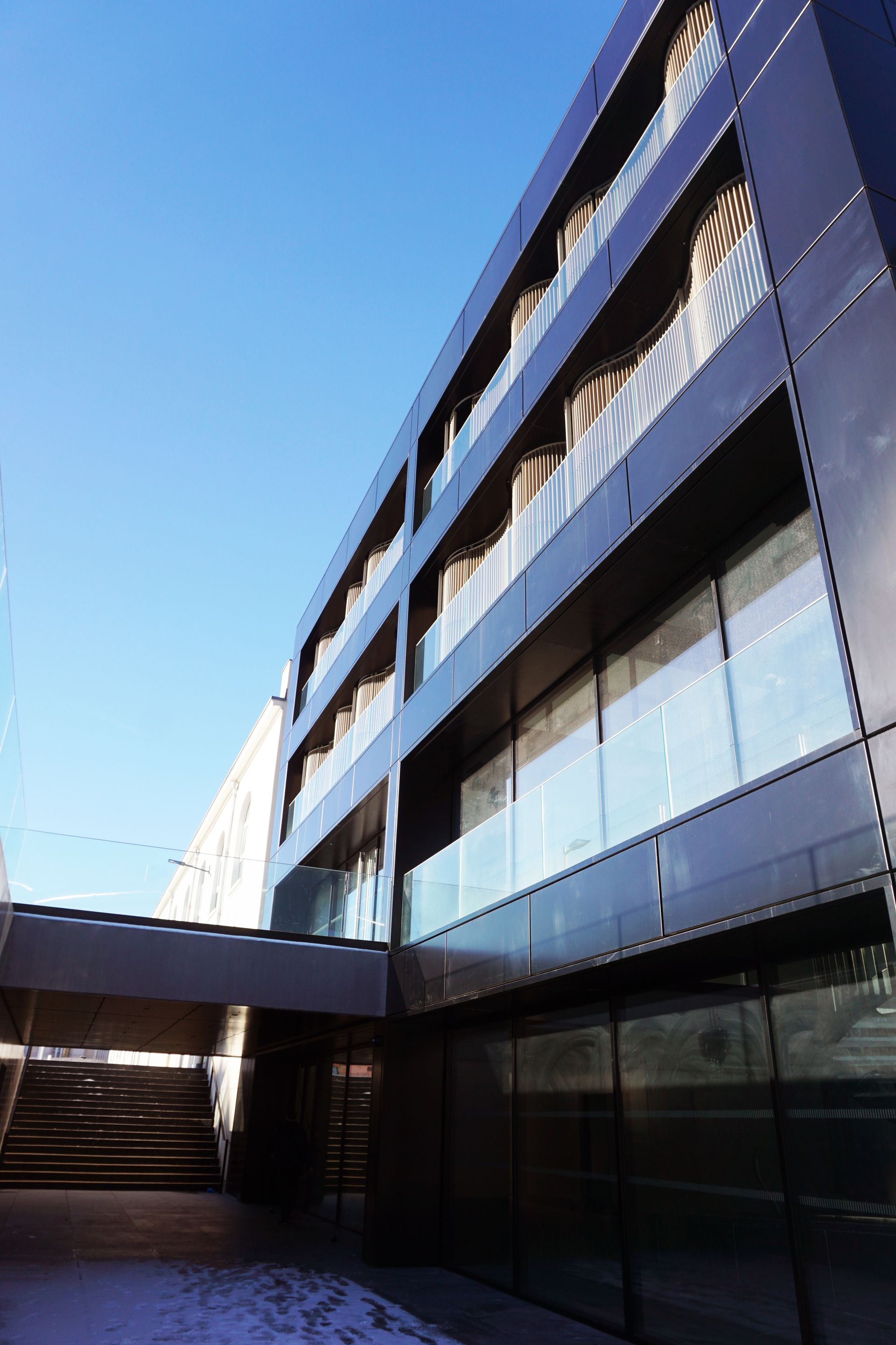 Bâtiment moderne et sombre avec balcons en verre et grandes fenêtres contre un ciel bleu.
