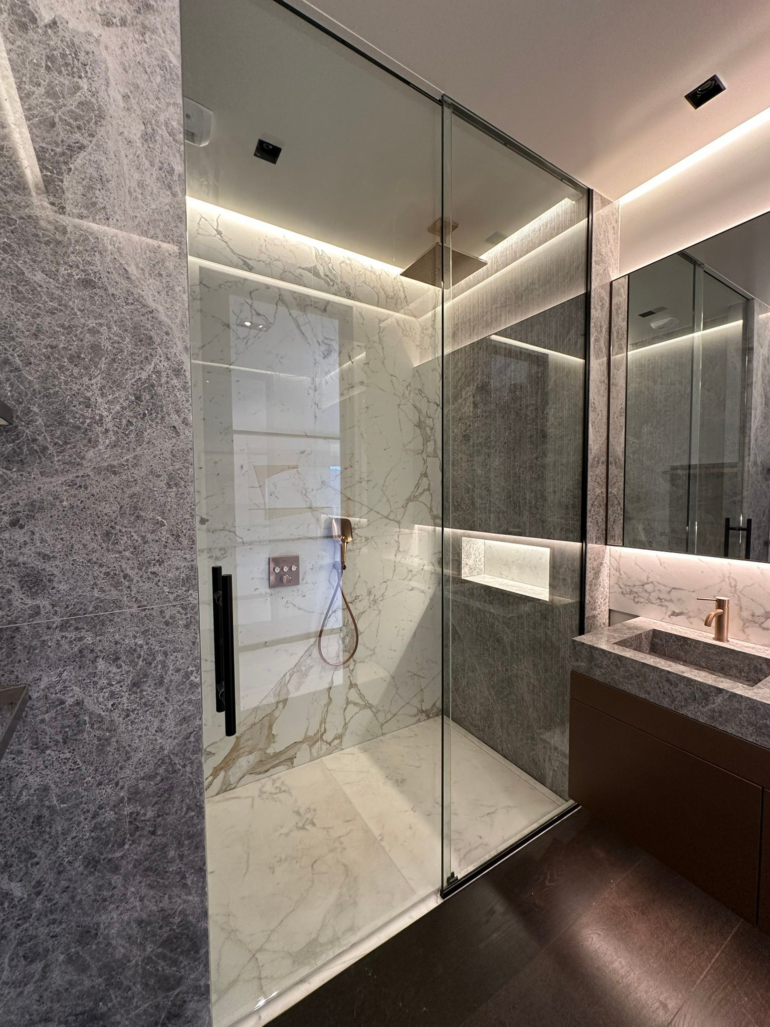 Salle de bain moderne avec douche en verre, murs en pierre grise, sol en carrelage de marbre et éclairage d'ambiance.
