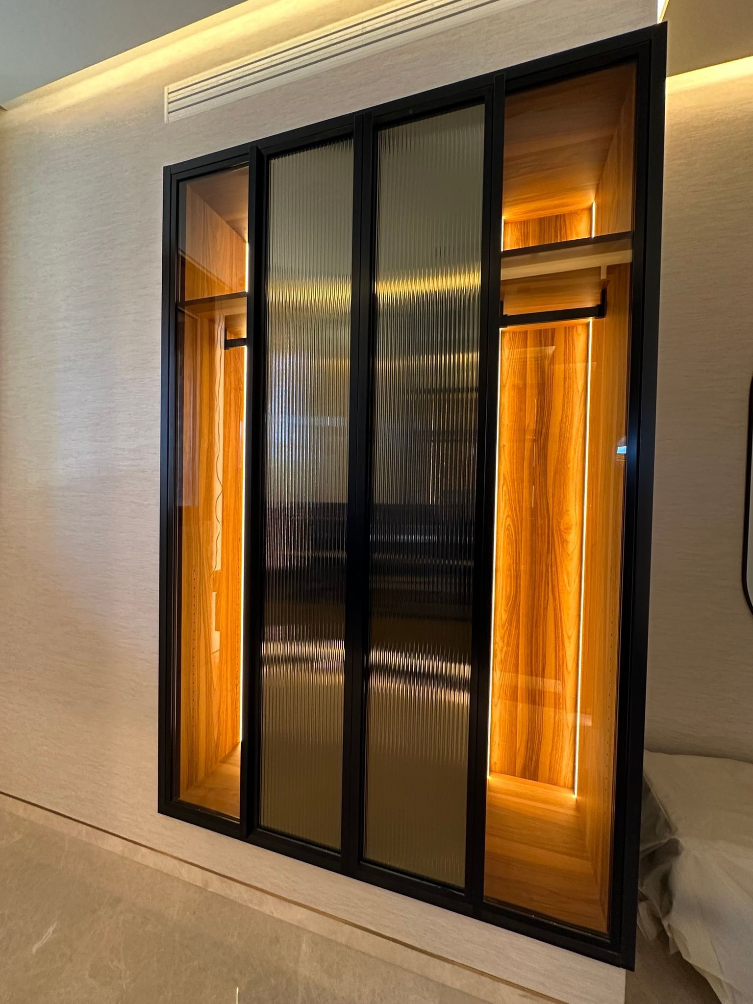 Placard à portes vitrées avec garniture noire, intérieur en bois éclairé et vêtements suspendus à l'intérieur.