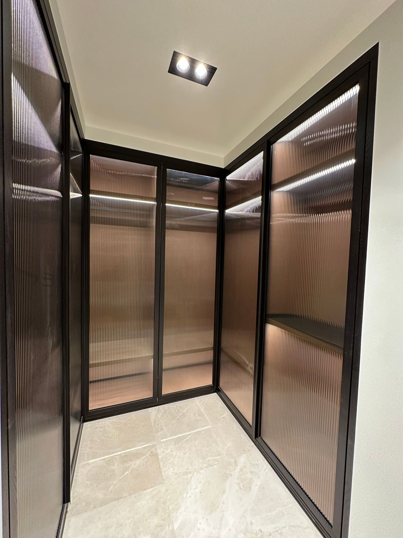 Dressing avec portes en verre ondulé couleur bronze, cadres foncés et sol neutre. Éclairage intérieur visible.