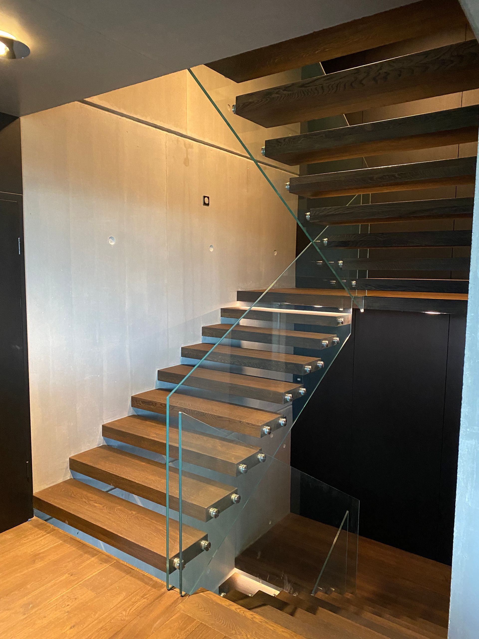 Escalier en bois avec rampe en verre, montant vers un plafond sombre.