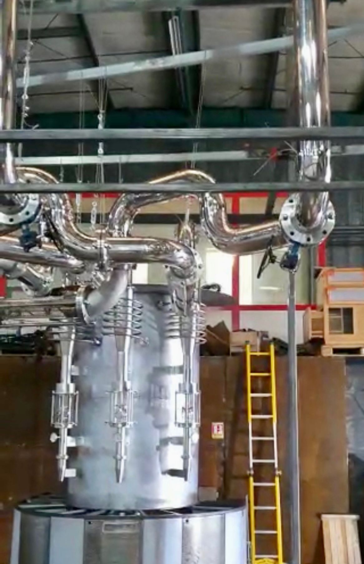 Grand réacteur industriel en acier inoxydable avec tuyauterie complexe, en configuration usine, avec une échelle jaune.
