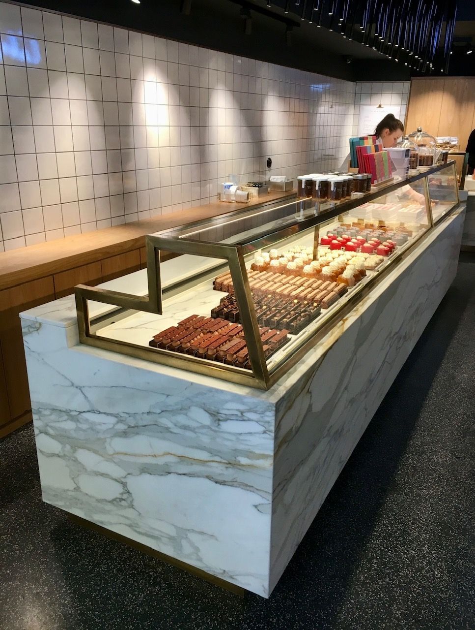 Comptoir de pâtisserie avec façade en marbre. Femme derrière ; pâtisseries à l'intérieur. Mur carrelé en arrière-plan.