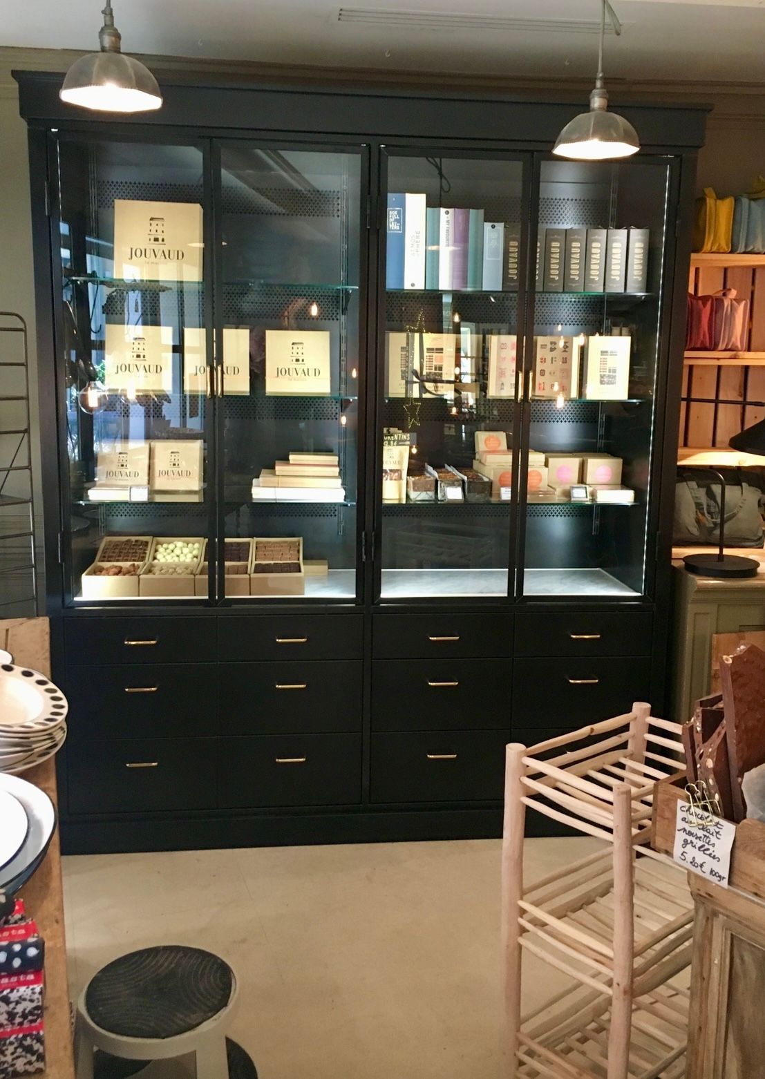 Vitrine noire avec portes vitrées, remplie de diverses boîtes et livres, éclairée par des lampes suspendues.