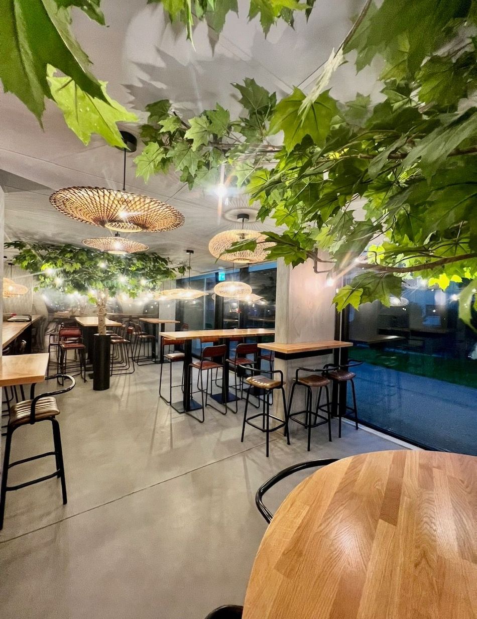 Restaurant intérieur avec tables en bois, tabourets noirs et suspensions. Végétation artificielle au plafond.
