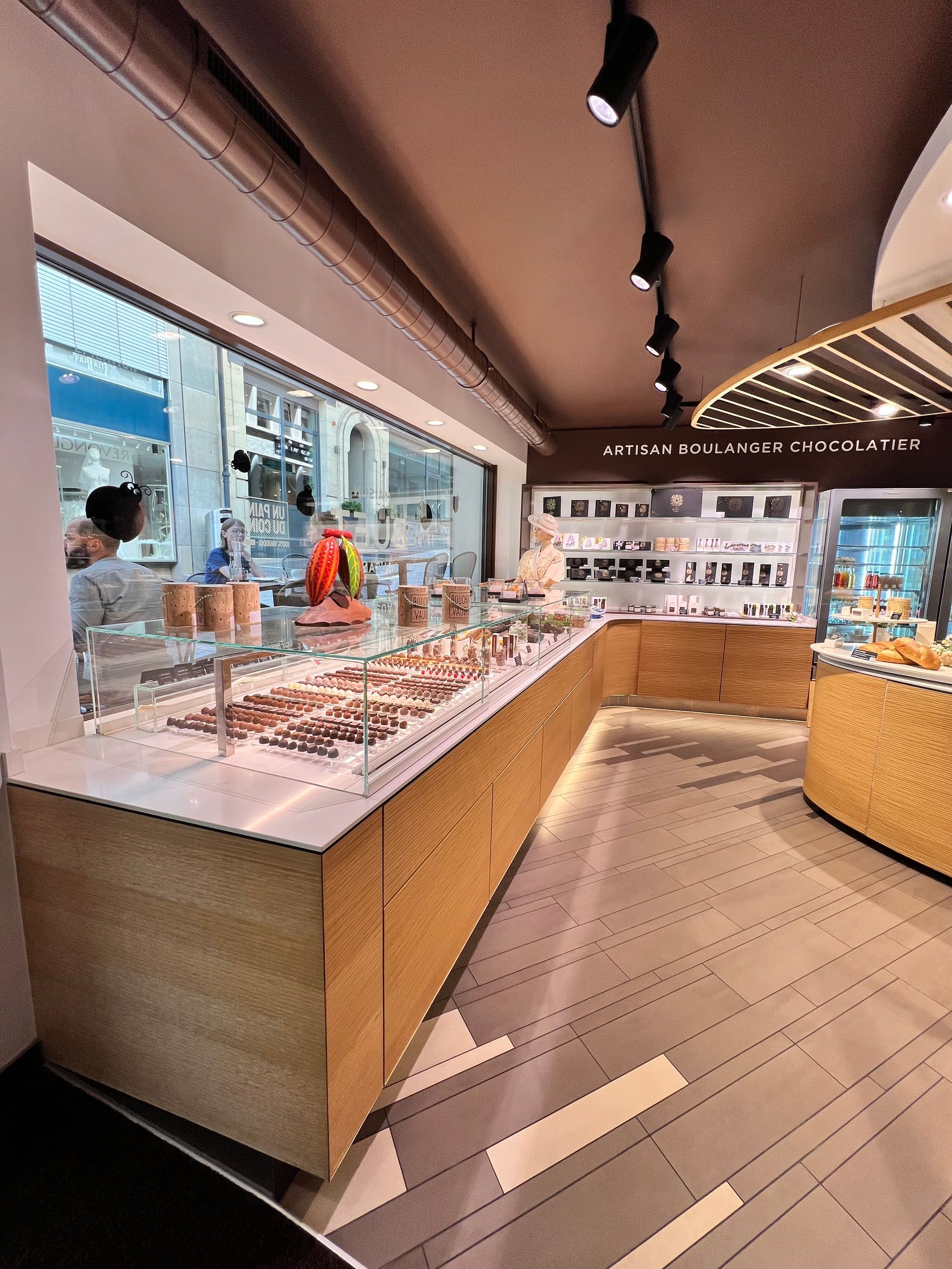 Une boulangerie avec un étalage de chocolats, des comptoirs en bois clair et une personne visible à travers la vitrine.