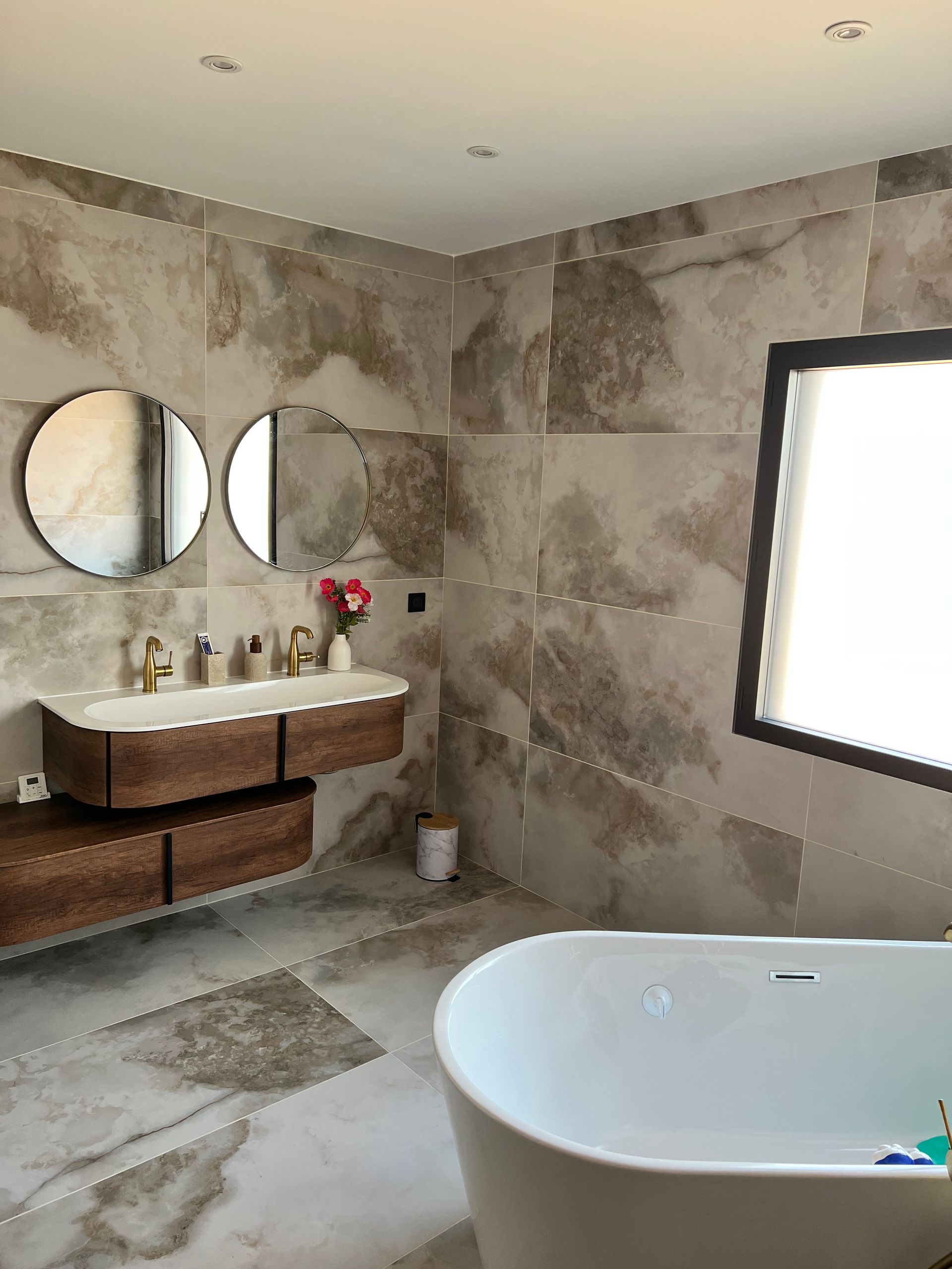 Salle de bain moderne avec carrelage aspect marbre, vanité flottante en bois, miroirs ronds et baignoire autonome.