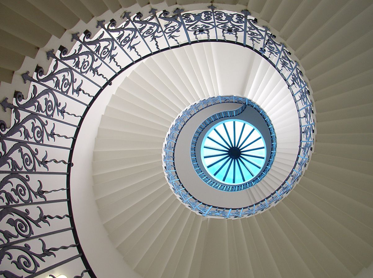 Escalier en colimaçon, vue vers le haut ; marches blanches, rampe en fer forgé noir, puits de lumière bleu.