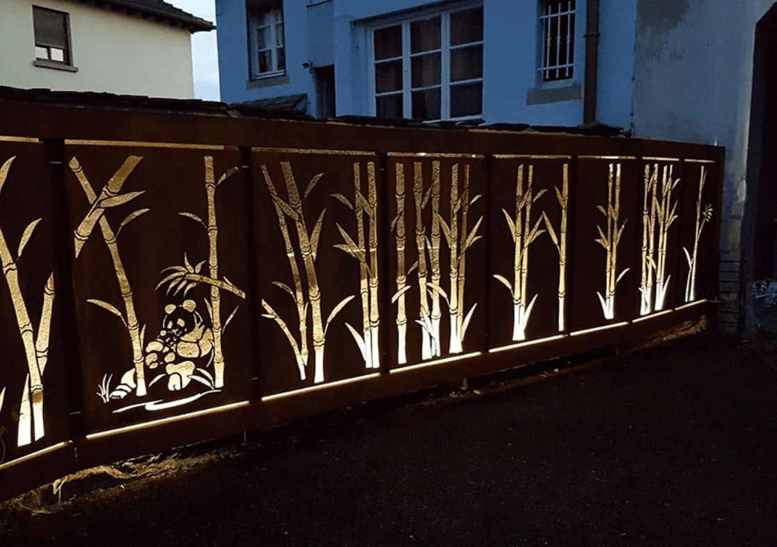 Clôture en métal rouillé avec un motif de silhouette en bambou, éclairée par l'arrière au crépuscule.