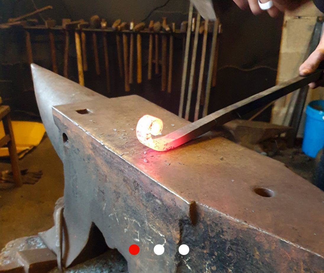 Un forgeron forge une spirale de métal rouge sur une enclume avec un marteau et des pinces dans un atelier.