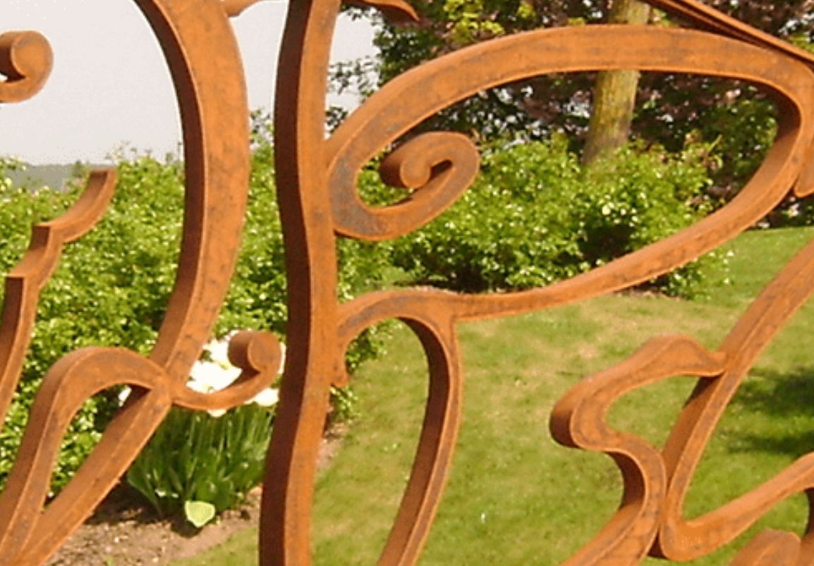 Clôture décorative en métal rouillé avec des tourbillons et des courbes stylisés, sur fond de pelouse verte et d'arbustes.