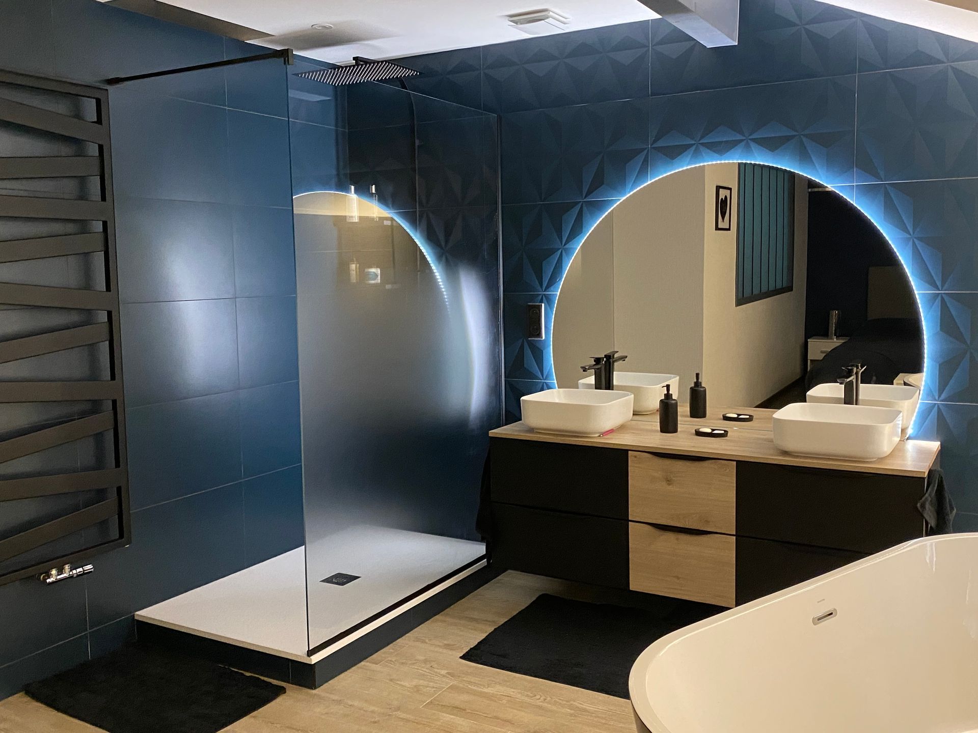 Salle de bain moderne avec murs bleus, douche à l'italienne, double vasque et miroir circulaire éclairé.