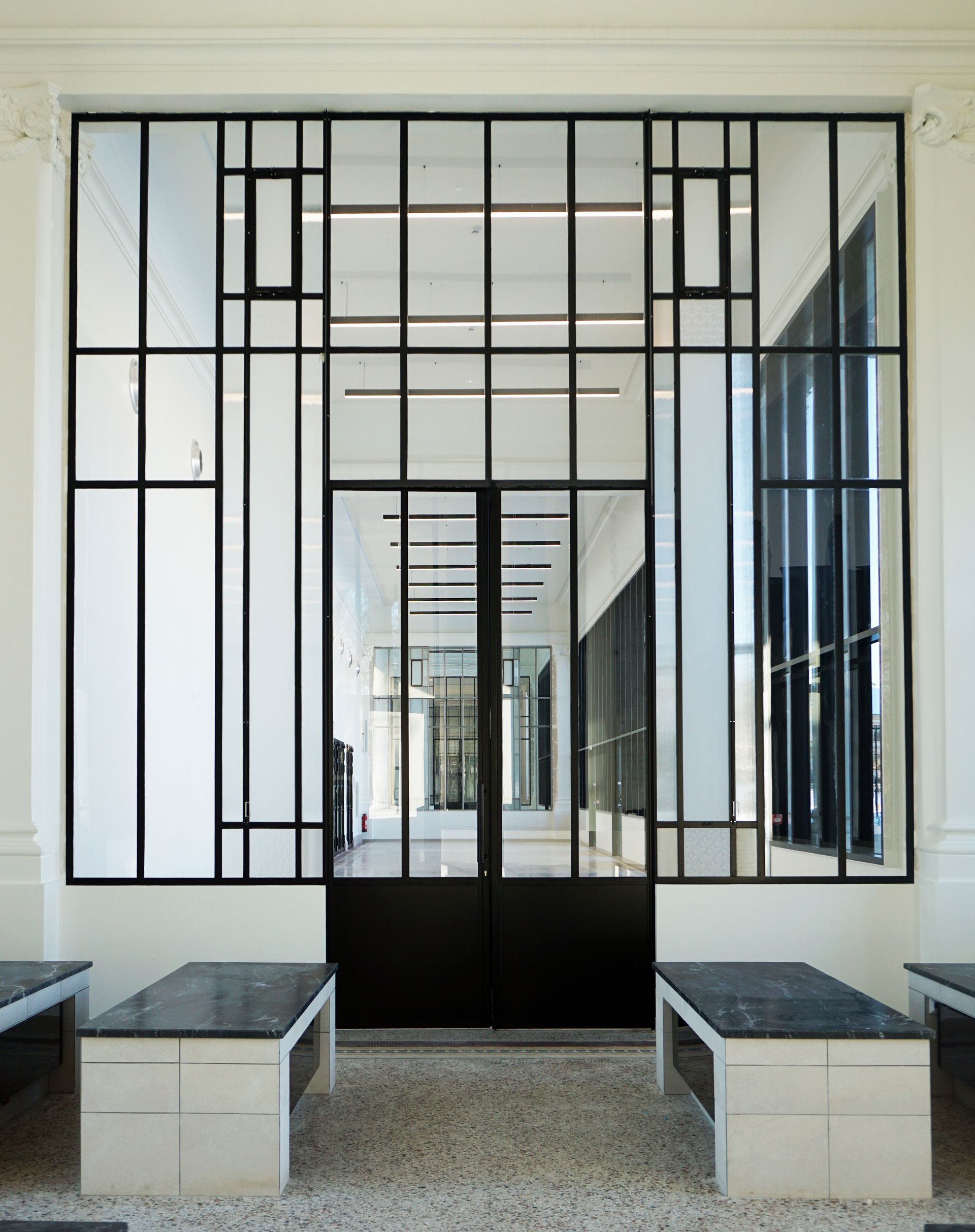 Porte en verre et miroir à cadre noir avec portes noires, murs blancs et bancs en marbre.
