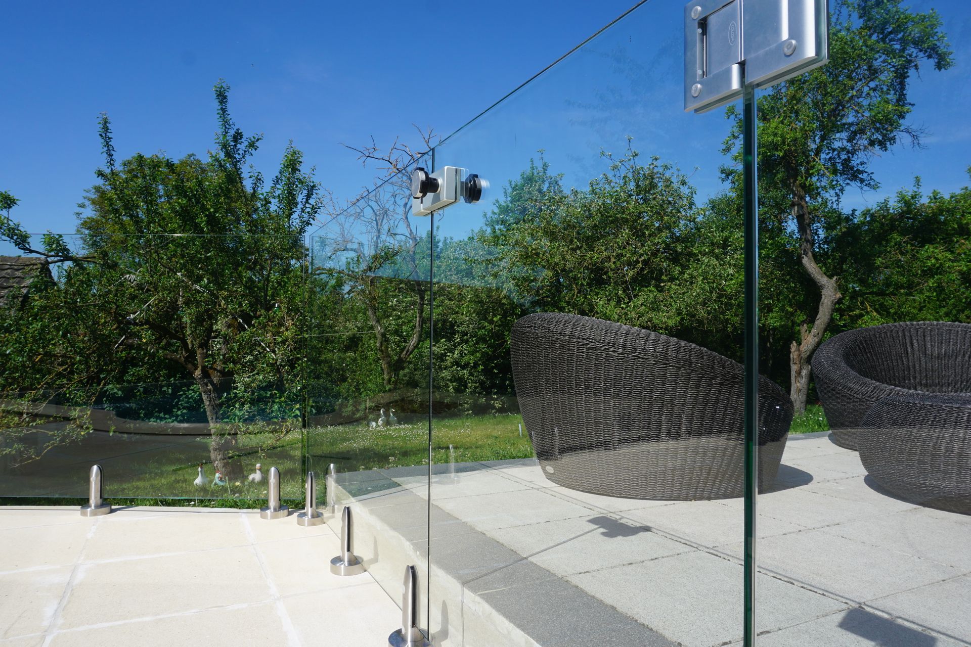 Clôture en verre donnant sur un patio avec des meubles en osier, des arbres et un ciel bleu clair.